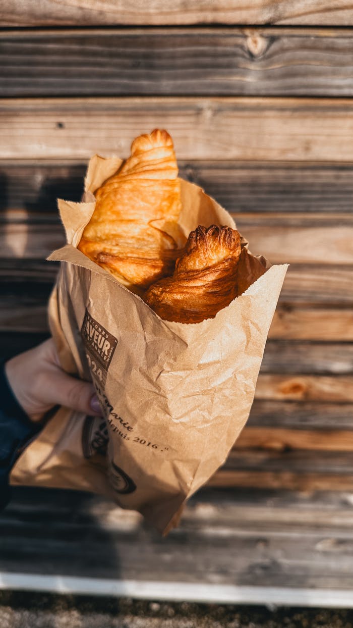 A person holding fresh croissants in a paper bag against a wooden background.
